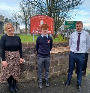 Grianghraf de bheirt múinteoirí agus scoláire os comhair comhartha scoile