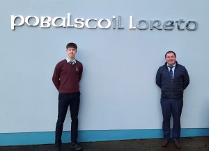 Grianghraf de scoláire agus múinteoir os comhair balla gorm a bhfuil ainm na scoile air